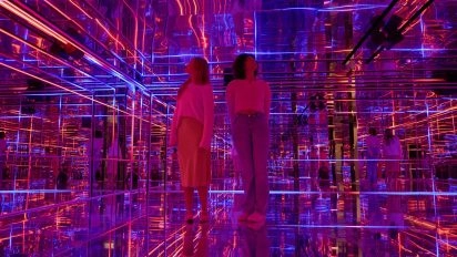 Two visitors inside a mirrored room filled with vibrant neon lights and geometric reflections by Pulse Interactive at Moco Museum London.