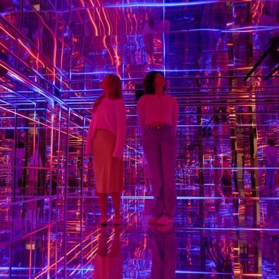 Dos visitantes dentro de una sala con espejos, luces de neón vibrantes y reflejos geométricos de Pulse Interactive en Moco Museum London.