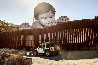 Large-scale installation by JR of a toddler peering over the US-Mexico border wall, with a border patrol vehicle below.