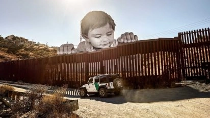 Grootschalige installatie van JR met een peuter die over de Amerikaans-Mexicaanse grensmuur kijkt, met een grenspatrouillevoertuig eronder.